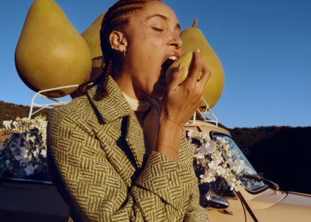 Adwoa Aboah con i capelli intrecciati addenta una pera sullo sfondo di tre grandi oggetti di scena a forma di pera posti sopra il portapacchi di un’auto.