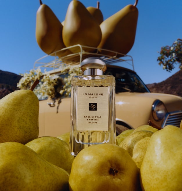 Flacone di English Pear & Freesia cologne da 100 ml circondata da pere fresche e sullo sfondo grandi oggetti di scena a forma di pera posti sopra il portapacchi di un’auto.