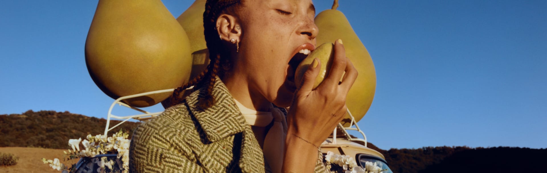 Adwoa Aboah, cheveux tressés, mordant dans une poire avec, en toile de fond, trois gros accessoires en forme de poire placés sur le toit d’une voiture.