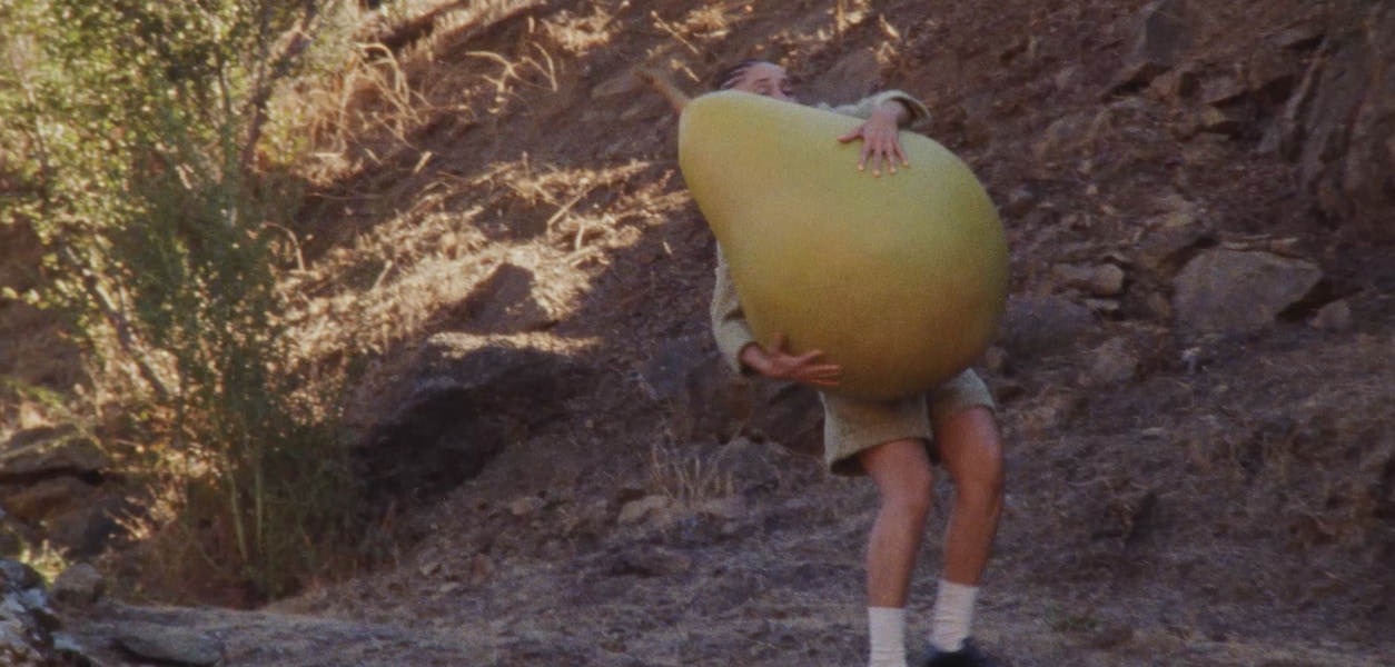 Vidéo d’Adwoa Aboah transportant un gros accessoire en forme de poire le long d’un chemin sec et rocailleux à flanc de colline.