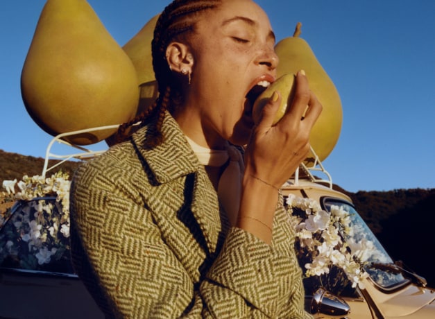 Adwoa Aboah, cheveux tressés, mordant dans une poire avec, en toile de fond, trois gros accessoires en forme de poire placés sur le toit d’une voiture.