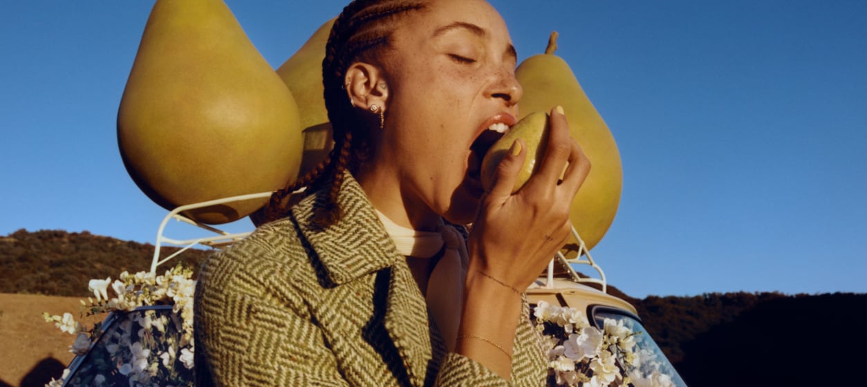 Adwoa Aboah con pelo trenzado, mordiendo una pera con un fondo de tres grandes peras de atrezo encima del portaequipajes de un coche.